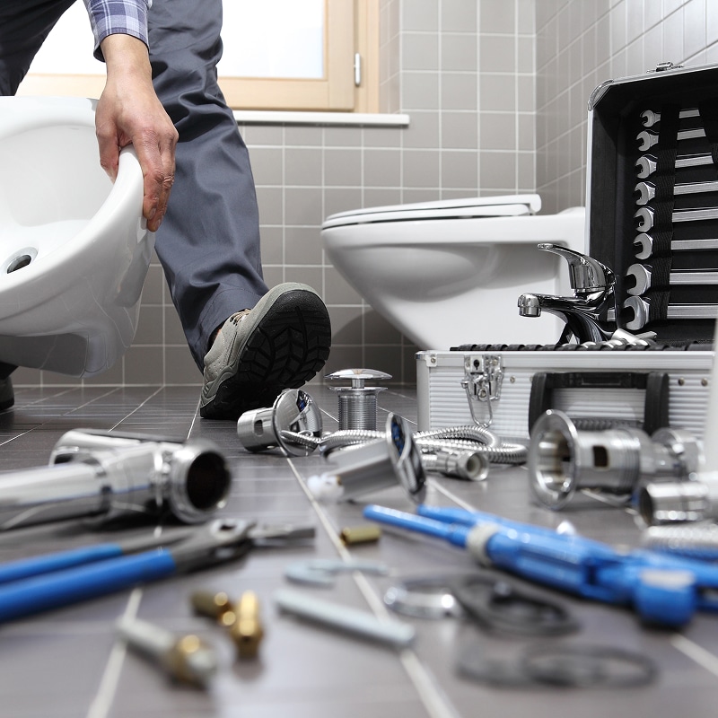 Plombier installant un bidet dans une salle de bain carrelée, avec outils, robinetterie et un WC en arrière-plan.