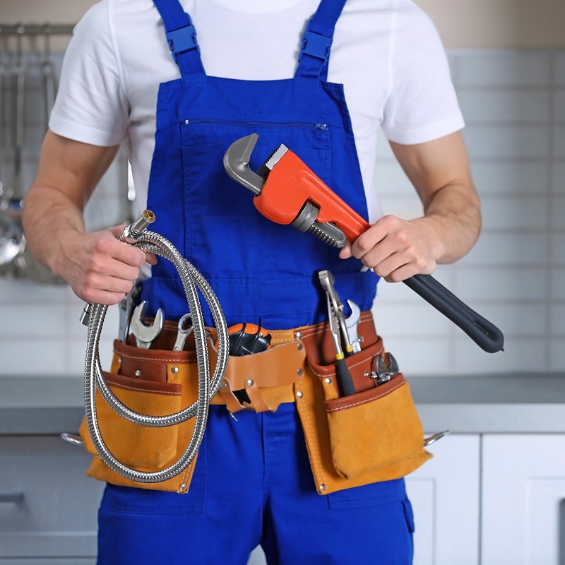 Plombier en tenue bleue, ceinture à outils garnie, tenant une clé à tube orange et un flexible métallique.