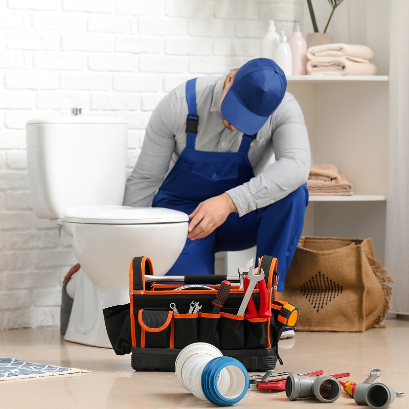 Plombier professionnel en combinaison bleue et casquette bleue répare une toilette blanche. Une boîte à outils remplie et des raccords de tuyauterie sont posés au sol.