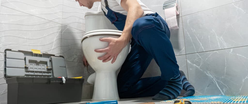 Plombier en combinaison bleue répare des toilettes blanches dans une salle de bain carrelée grise. Boîte à outils ouverte à côté.