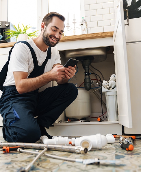 Plombier souriant consulte son téléphone sous un évier de cuisine. Il répare la plomberie, entouré de tuyaux et outils.