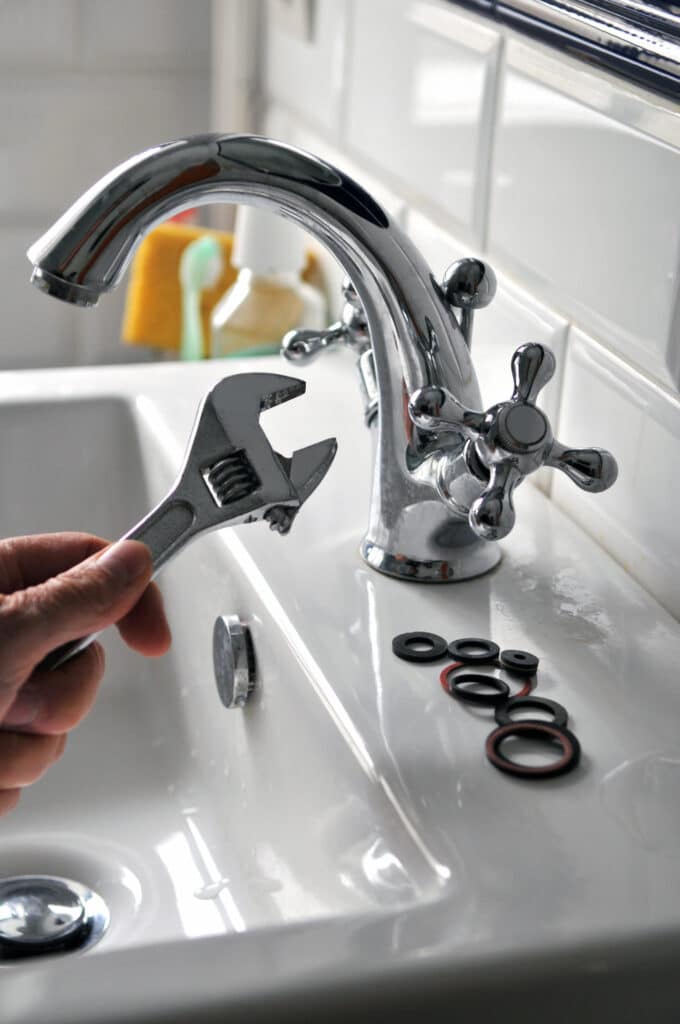 Main tenant une clé à molette, prête à réparer un robinet chromé sur un lavabo blanc. Joints d'étanchéité noirs et rouges à côté.