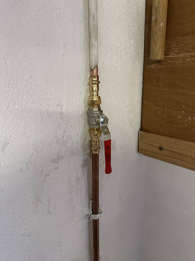 Conduite verticale murale: tube transparent, raccord laiton, tuyau cuivre avec valve à poignée rouge 'A'. Collier de fixation visible.