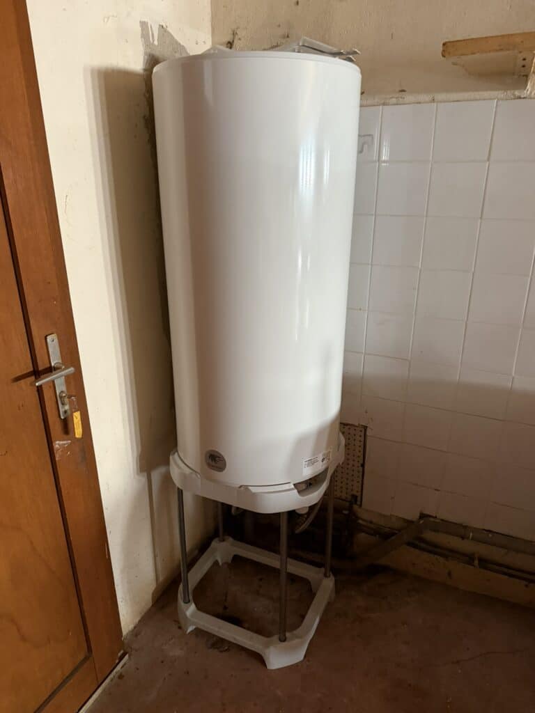Grand chauffe-eau Thermor blanc sur pied, installé dans pièce utilitaire avec mur carrelé et porte en bois. Une clé est dans la serrure.