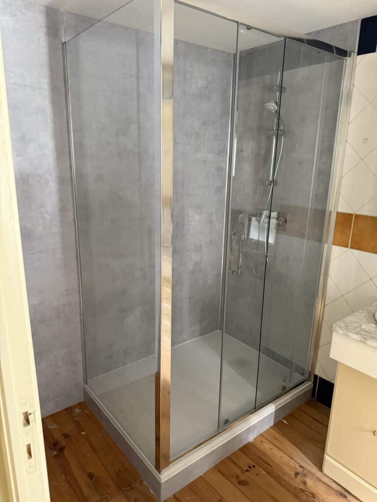 Cabine de douche moderne avec parois gris béton et verre, bac blanc, installée sur un plancher en bois. Meuble vasque à droite.