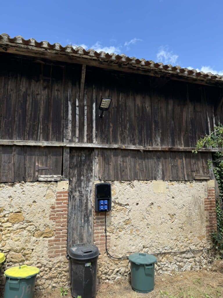 Mur ancien de grange en bois et pierre, tuiles, éclairage LED solaire et boîtier électrique. Deux poubelles au sol.