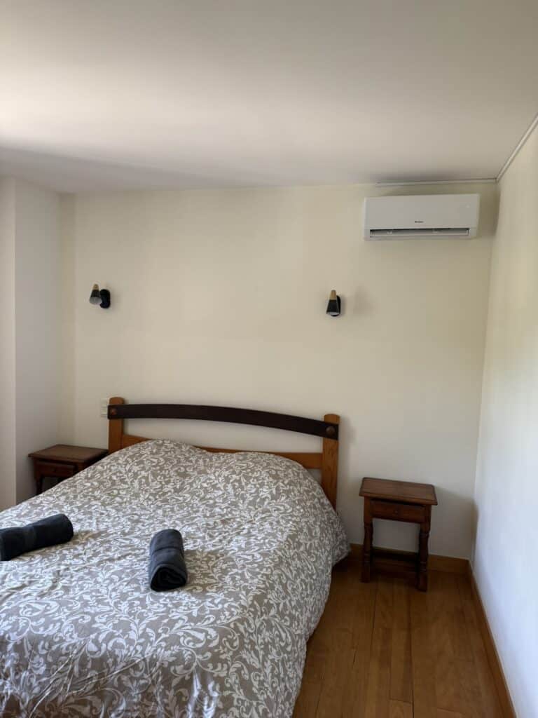 Chambre double lumineuse avec lit à motif floral, deux chevets en bois, appliques murales, climatiseur Gree et parquet.
