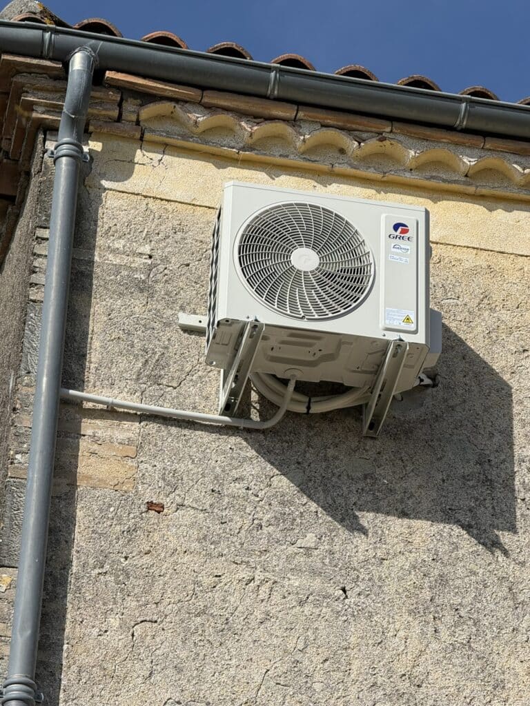 Unité Extérieure Climatisation GREE Inverter Murale Unité de climatisation Gree Inverter sur un vieux mur en pierre texturé, sous un toit de tuiles et une gouttière grise. Des fissures parcourent le mur.