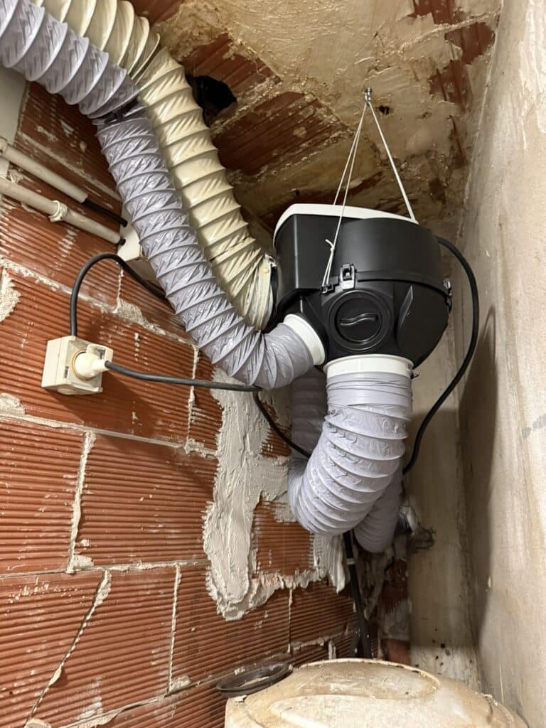 Unité de ventilation noire suspendue, avec conduits souples gris et beiges connectés, dans un local aux murs de briques. Prise électrique visible.